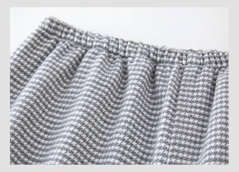 May include: Close-up of grey and white houndstooth fabric, possibly from trousers or shorts. An elasticated waistband is visible at the top, with the fabric gathered below. The material has a soft, textured appearance.