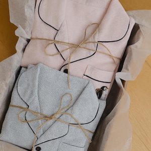 May include: Two sets of pajamas, one pink and one gray, are neatly folded in a box lined with tissue paper. The pajamas have black piping and are tied with twine. A small penguin embroidery is visible on the gray set.
