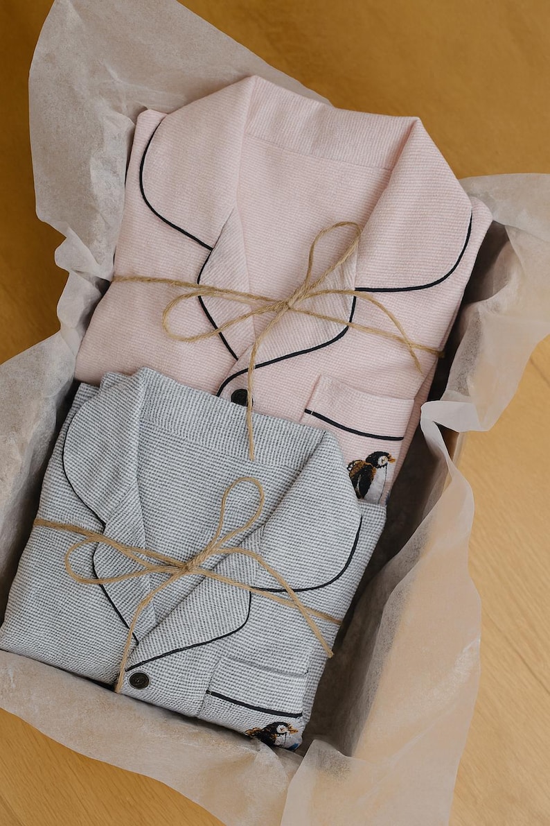May include: Two sets of pajamas, one pink and one gray, are neatly folded in a box lined with tissue paper. The pajamas have black piping and are tied with twine. A small penguin embroidery is visible on the gray set.