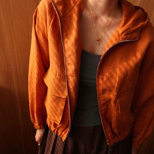 May include: An orange hooded jacket with a full zipper closure and two front pockets. The jacket is made of a textured fabric and has long sleeves. The model is wearing a gray tank top and brown pants. A gold necklace is visible.