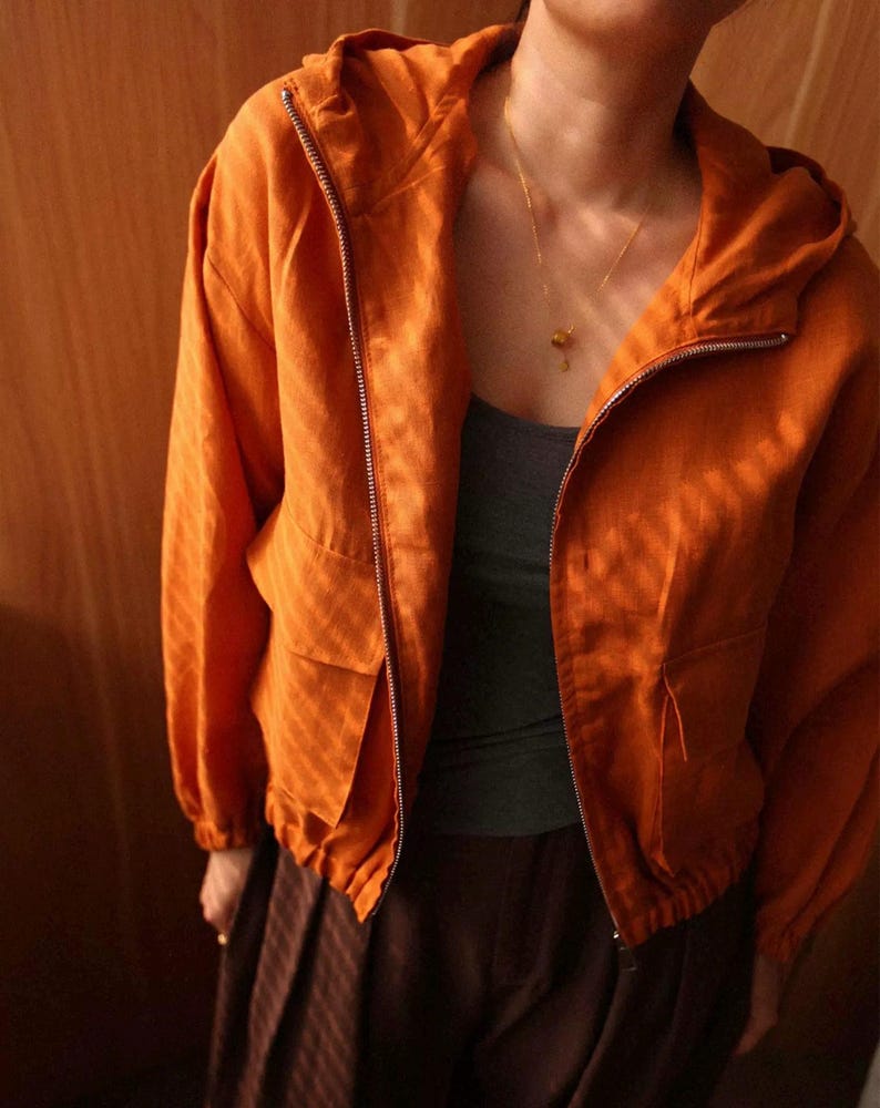 May include: An orange hooded jacket with a full zipper closure and two front pockets. The jacket is made of a textured fabric and has long sleeves. The model is wearing a gray tank top and brown pants. A gold necklace is visible.