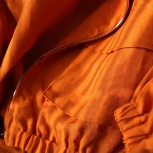 May include: Close-up of an orange jacket made of a textured fabric. The jacket features a silver zipper, a pocket, and gathered cuffs. The fabric has visible folds and creases, suggesting a lightweight material.
