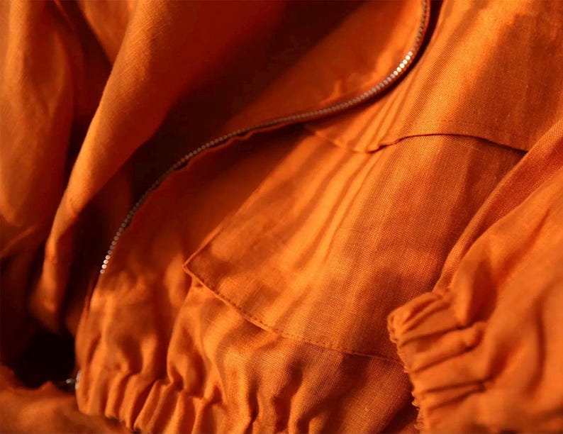 May include: Close-up of an orange jacket made of a textured fabric. The jacket features a silver zipper, a pocket, and gathered cuffs. The fabric has visible folds and creases, suggesting a lightweight material.