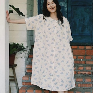 Puede incluir: Vestido crema con botones, mangas cortas y estampado floral azul claro. El vestido llega a la pantorrilla y tiene un corte holgado. El vestido está hecho de una tela ligera.