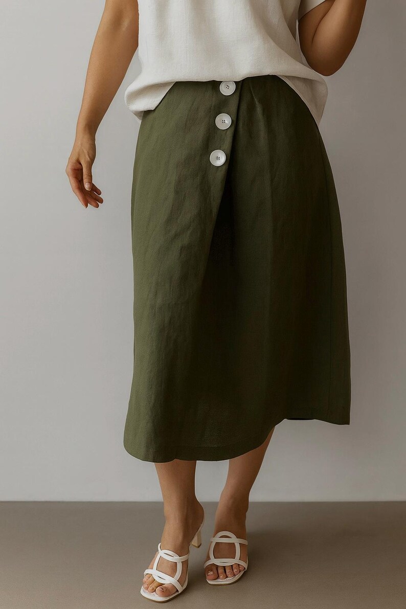 Pu&ograve; includere: Gonna a portafoglio verde oliva con tre grandi bottoni bianchi. La gonna &egrave; lunga fino al ginocchio ed &egrave; realizzata in un tessuto fluido. La modella indossa sandali bianchi con un piccolo tacco.