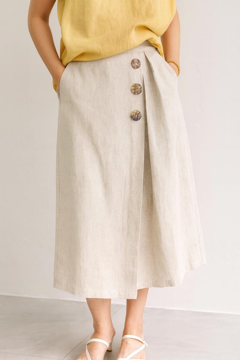 Pu&ograve; includere: Gonna in lino beige con parte anteriore a portafoglio, con tre grandi bottoni rotondi. La gonna ha la vita alta e arriva a met&agrave; polpaccio. La modella indossa un top giallo e la gonna ha tasche.