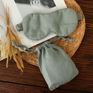 May include: A sage green sleep mask and matching drawstring pouch. The mask has an elastic band and is made of a soft, textured fabric. The pouch is closed with a drawstring. The items are displayed on a woven mat.