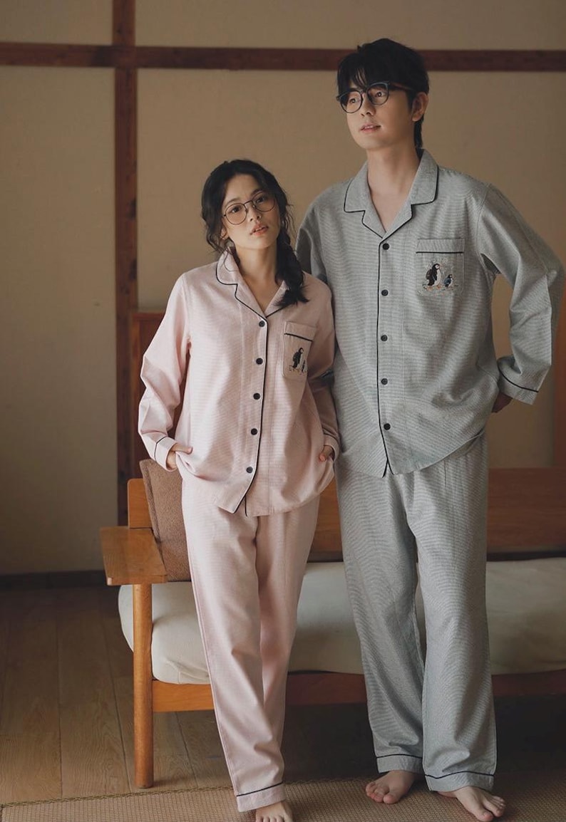May include: Matching pajama sets in pink and gray. The pink set has a light stripe pattern, black trim, and a penguin graphic. The gray set has a similar design. Both sets include a button-down top and pants.