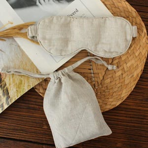 May include: A beige linen sleep mask and matching drawstring pouch. The mask has a curved shape to fit the eyes and is designed to block out light. The pouch is small and can be used to store the mask. The items are made of a natural-looking fabric.