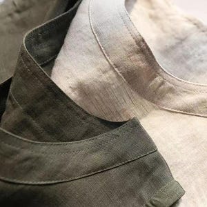 May include: Close-up of two linen shirts. One shirt is olive green, and the other is a light beige. The collars and necklines are visible, showcasing the texture and weave of the fabric. The shirts appear to be new.