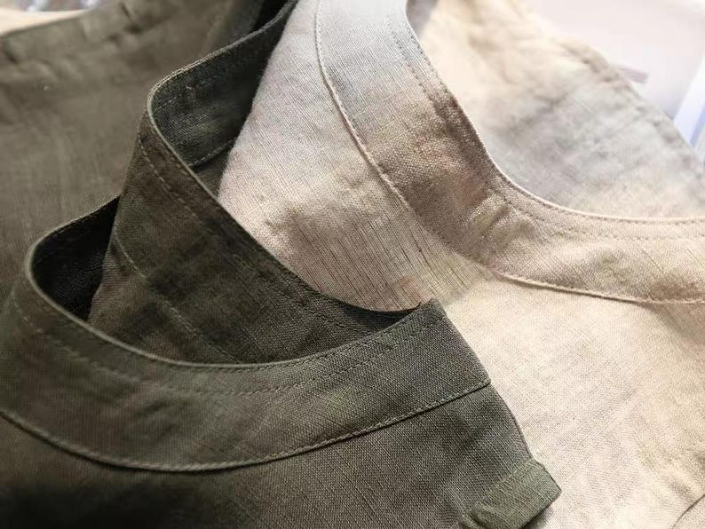 May include: Close-up of two linen shirts. One shirt is olive green, and the other is a light beige. The collars and necklines are visible, showcasing the texture and weave of the fabric. The shirts appear to be new.