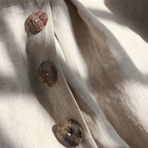 Pu&ograve; includere: Primo piano di un capo in lino beige con tre grandi bottoni rotondi. I bottoni sono un mix di marrone e beige, con un aspetto naturale e organico. Il tessuto ha una trama sottile e un colore morbido e neutro.