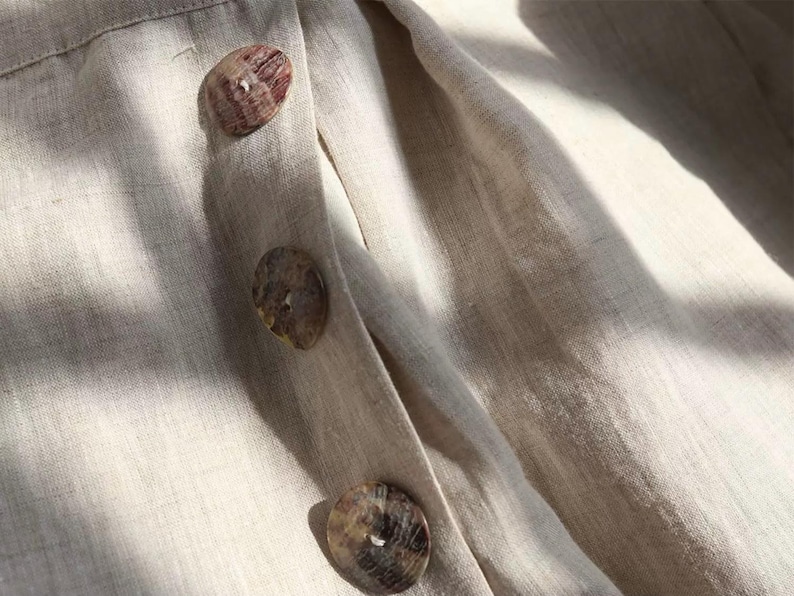 Pu&ograve; includere: Primo piano di un capo in lino beige con tre grandi bottoni rotondi. I bottoni sono un mix di marrone e beige, con un aspetto naturale e organico. Il tessuto ha una trama sottile e un colore morbido e neutro.