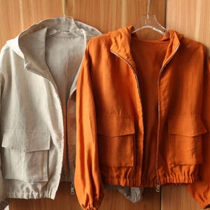 May include: Two hooded jackets hanging on a wooden background. One jacket is a light beige color, and the other is a vibrant orange. Both jackets have a zipper closure, two front pockets, and a drawstring hem. The jackets are made of a lightweight fabric.