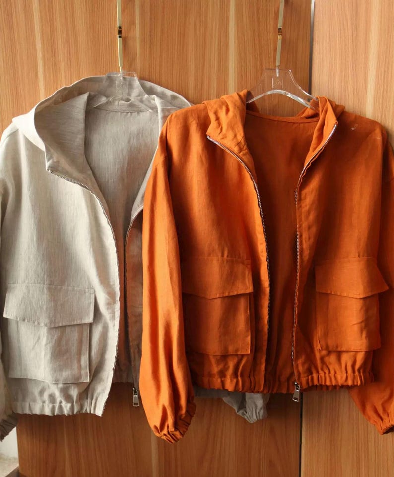 May include: Two hooded jackets hanging on a wooden background. One jacket is a light beige color, and the other is a vibrant orange. Both jackets have a zipper closure, two front pockets, and a drawstring hem. The jackets are made of a lightweight fabric.