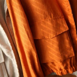 May include: Close-up of two jackets. One is a light beige color, and the other is a vibrant orange. The orange jacket has two front pockets and a zipper closure. Both jackets appear to be made of a lightweight fabric.