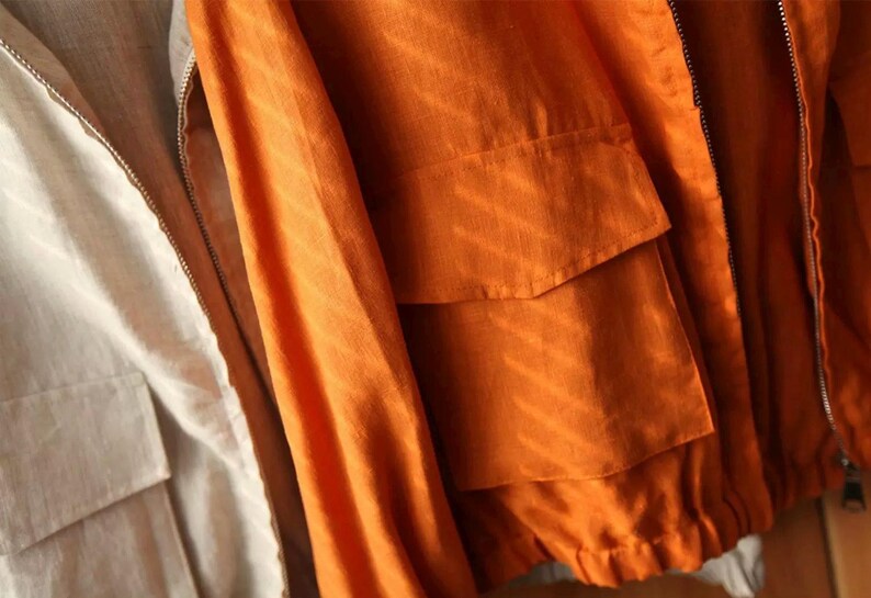 May include: Close-up of two jackets. One is a light beige color, and the other is a vibrant orange. The orange jacket has two front pockets and a zipper closure. Both jackets appear to be made of a lightweight fabric.
