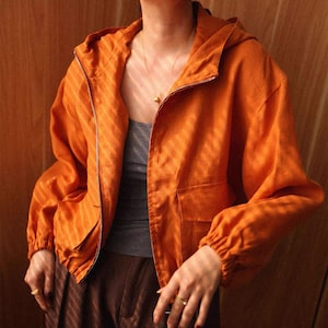 Puede incluir: Chaqueta naranja con capucha, cierre de cremallera y dos bolsillos delanteros. La chaqueta tiene mangas largas con puños elásticos. La chaqueta se lleva sobre una camiseta sin mangas gris y pantalones marrones. Se ve un collar dorado.