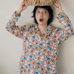 May include: A beige pajama set with a repeating pattern of cartoon bears and blue accents. The long-sleeved button-down shirt and matching pants are worn by a person holding a woven basket above their head.