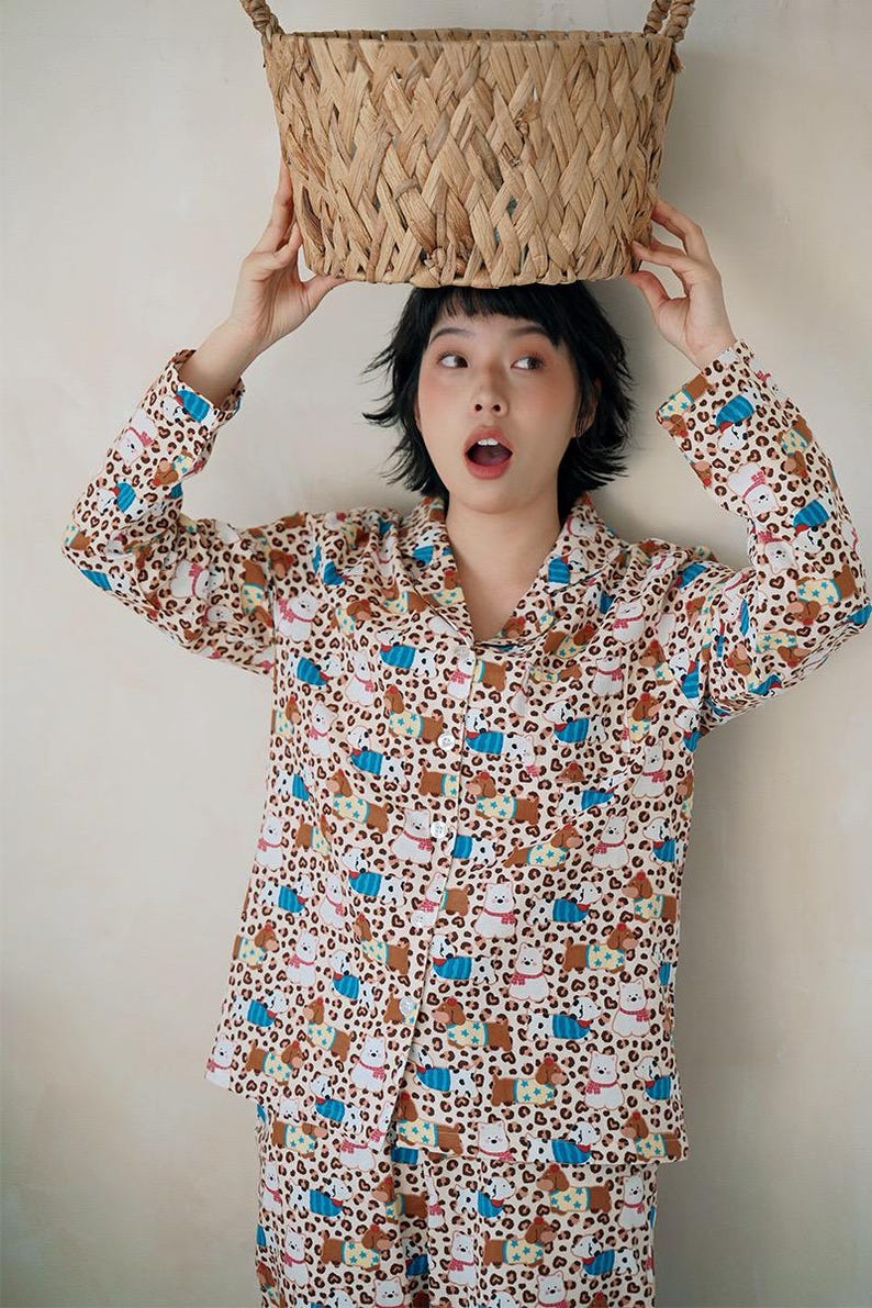 May include: A beige pajama set with a repeating pattern of cartoon bears and blue accents. The long-sleeved button-down shirt and matching pants are worn by a person holding a woven basket above their head.