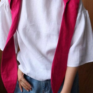May include: A white linen top with short sleeves and a round neckline. A bright pink scarf is draped over the shoulders. The person is wearing blue jeans.