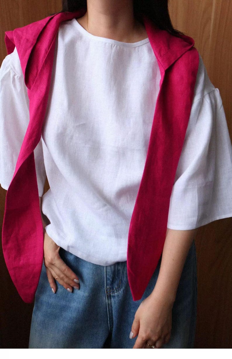 May include: A white linen top with short sleeves and a round neckline. A bright pink scarf is draped over the shoulders. The person is wearing blue jeans.