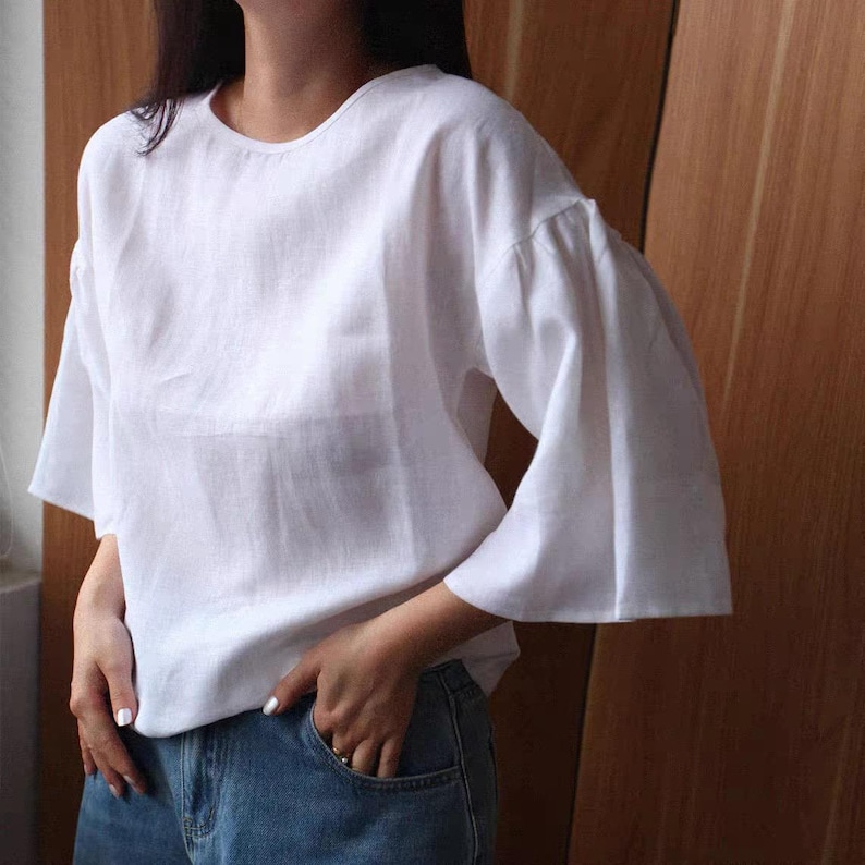 May include: A white linen top with a round neckline and flared sleeves. The top is a loose fit and appears to be made of a lightweight fabric. The sleeves are a distinctive feature, adding a touch of elegance to the simple design. The model is wearing blue jeans.