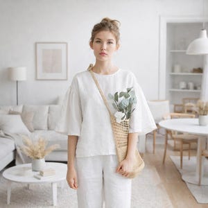 May include: A person wearing a white linen top and pants, holding a beige net bag filled with greenery and white flowers. The outfit is simple and elegant. The background shows a bright, minimalist living space with white furniture and decor.