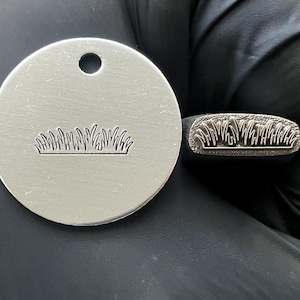 Puede incluir: Una etiqueta redonda de metal plateado con un diseño de hierba y un agujero en la parte superior. Un sello de metal con el mismo diseño de hierba está al lado de la etiqueta. El diseño de la hierba es una serie de líneas curvas.