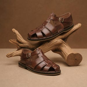 Op de afbeelding: Twee bruine leren sandalen met een kooi-ontwerp en verstelbare gespen. De sandalen worden gepresenteerd op een stuk drijfhout tegen een neutrale achtergrond. De zolen hebben een zwarte rand met witte stiksels.