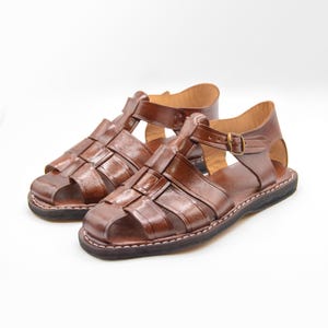 Op de afbeelding: Een paar bruine leren sandalen met een geweven ontwerp over de bovenkant. De sandalen hebben verstelbare enkelbandjes met een metalen gesp en een gestikte zool. Op een witte achtergrond.