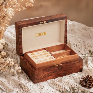 Peut inclure: Une boîte à bijoux rectangulaire en bois avec une finition en loupe sombre et un couvercle à charnière. L'intérieur présente une doublure en velours crème avec le texte doré Enom sur le couvercle et un plateau amovible contenant des bagues.