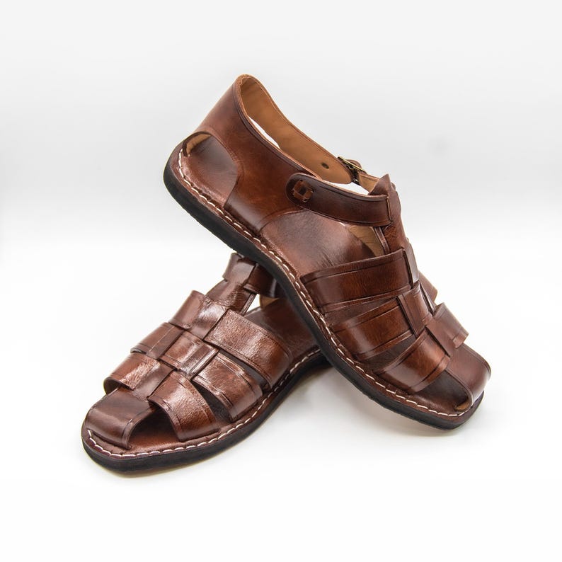 Op de afbeelding: Een paar bruin lederen sandalen met een geweven ontwerp aan de voorkant. De sandalen hebben een gesloten neus, een enkelband en een zwarte zool met witte stiksels. De sandalen worden op een witte achtergrond gepresenteerd.