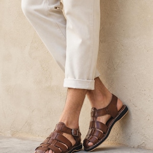Op de afbeelding: Bruine leren sandalen met een geweven design en een gespsluiting. De sandalen hebben een zwarte zool en worden gedragen met een beige broek. Klassieke stijl.