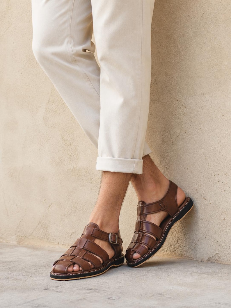 Op de afbeelding: Bruine leren sandalen met een geweven design en een gespsluiting. De sandalen hebben een zwarte zool en worden gedragen met een beige broek. Klassieke stijl.