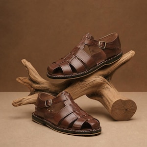 Op de afbeelding: Twee bruine leren sandalen met een kooi-achtig ontwerp en verstelbare gespen. De sandalen worden gepresenteerd op een stuk drijfhout tegen een bruine achtergrond. De zolen hebben een zwarte rand met witte stiksels.