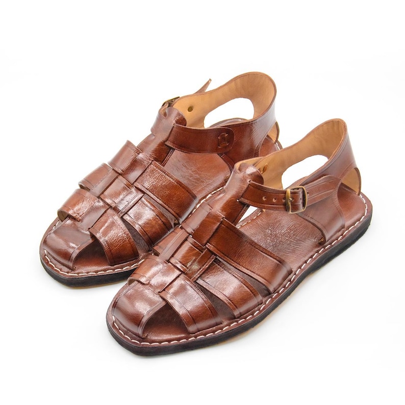 Op de afbeelding: Een paar bruinleren sandalen met een geweven ontwerp. De sandalen hebben verstelbare enkelbanden met metalen gespen en zijn aan de randen gestikt. Gepresenteerd op een witte achtergrond.