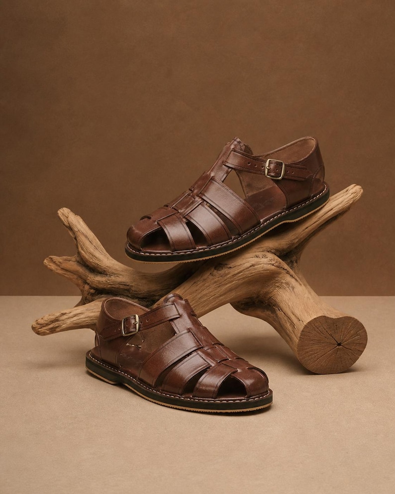 Op de afbeelding: Twee bruine leren sandalen met een kooi-ontwerp en verstelbare gespen. De sandalen worden gepresenteerd op een stuk drijfhout tegen een neutrale achtergrond. De zolen hebben een zwarte rand met witte stiksels.
