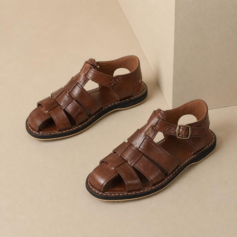 Op de afbeelding: Een paar bruin lederen sandalen met een geweven design. De sandalen hebben een gesloten teenontwerp met meerdere bandjes en een gespsluiting. De zolen zijn zwart met een wit stikseldetail. De sandalen zijn ongeveer 25 cm lang.