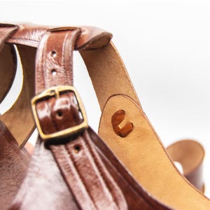 Op de afbeelding: Close-up van een bruine leren sandaal met een gesp en bandjes. De sandaal heeft een geweven ontwerp en een messingkleurige gesp. Het leer lijkt versleten, met zichtbare stiksels en een natuurlijke afwerking.