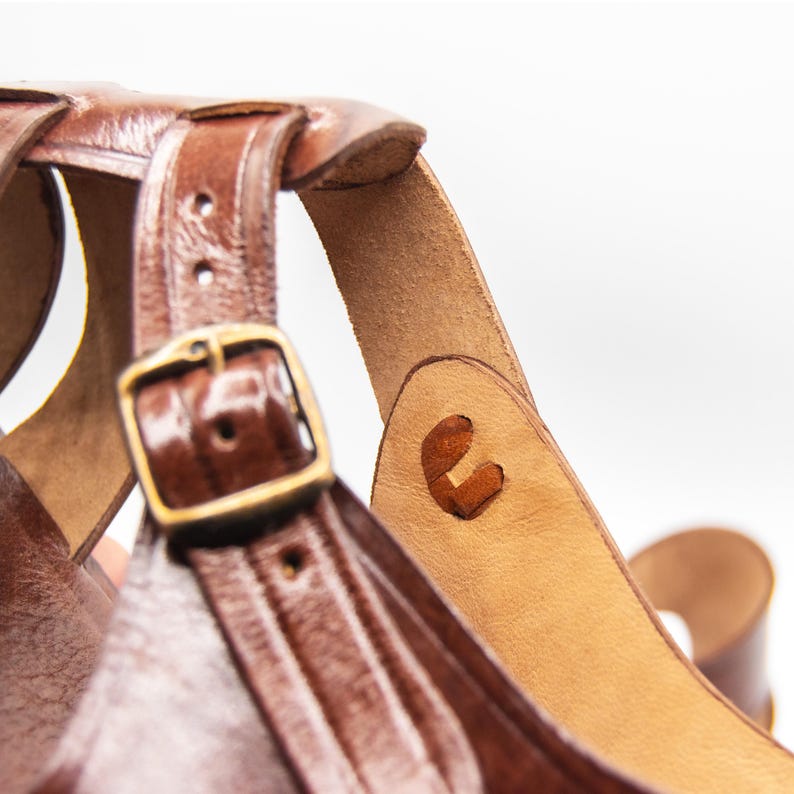 Op de afbeelding: Close-up van een bruine leren sandaal met een gesp en bandjes. De sandaal heeft een geweven ontwerp en een messingkleurige gesp. Het leer lijkt versleten, met zichtbare stiksels en een natuurlijke afwerking.