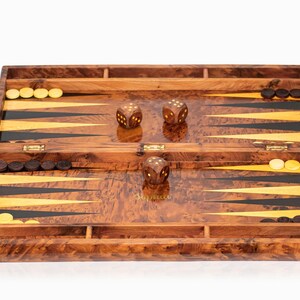 Pu&ograve; includere: Un set da backgammon in legno con finitura marrone e dorata. Il tabellone &egrave; aperto e mostra la superficie di gioco con triangoli neri e dorati. Ci sono pedine bianche e nere e dadi sul tabellone.