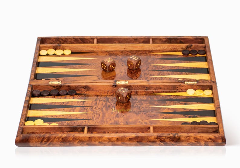 Pu&ograve; includere: Un set da backgammon in legno con finitura marrone e dorata. Il tabellone &egrave; aperto e mostra la superficie di gioco con triangoli neri e dorati. Ci sono pedine bianche e nere e dadi sul tabellone.