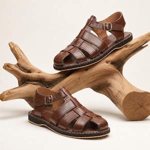 Puede incluir: Dos sandalias de cuero marrón con un diseño tejido y correas de hebilla ajustables. Las sandalias se exhiben sobre un trozo de madera flotante sobre un fondo neutro. Las sandalias tienen una suela negra.
