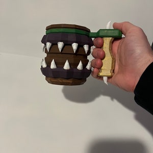 May include: A handcrafted mug with a barrel design. The mug has a brown and purple body with white, pointed teeth around the middle. The handle is gold and green. The mug is held by a person.