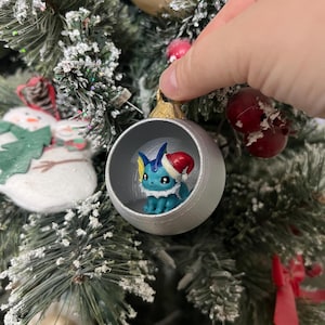 May include: A spherical Christmas ornament featuring a small blue Vaporeon figure wearing a red and white Santa hat inside the hollow center. The ornament has a matte silver exterior finish and a gold-colored cap at the top for hanging.