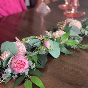 May include: A pink and green floral garland with eucalyptus leaves and pink flowers. The garland is on a wooden table.
