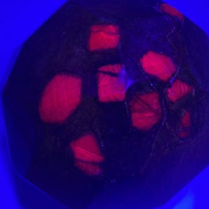 May include: A dark red gemstone with a faceted, irregular shape. The gemstone is illuminated with a UV light, revealing a pattern of bright red crystals inside.