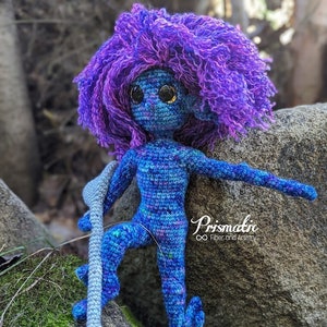 Könnte beinhalten: Eine gehäkelte Puppe in Blau und Lila mit großen Augen und einem langen, grauen Stock in der Hand. Die Puppe steht auf einem Stein mit grünem Moos.