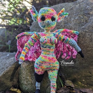 May include: A colorful crocheted amigurumi figure with wings, standing on a rock. The figure has a rainbow-colored body, black eyes, and black legs. The wings are pink and have a layered design. The figure is standing on a gray rock with green foliage in the background. The text "Prismatic Fiber and Artistry" is visible in the lower right corner of the image.